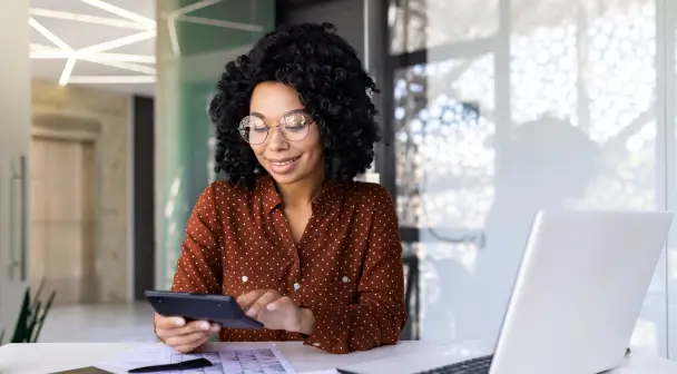 Eine Frau überprüft lächelnd in einem Büro, umgeben von ihrem Laptop und Unterlagen, ihre Versicherung online über ihr Handy.